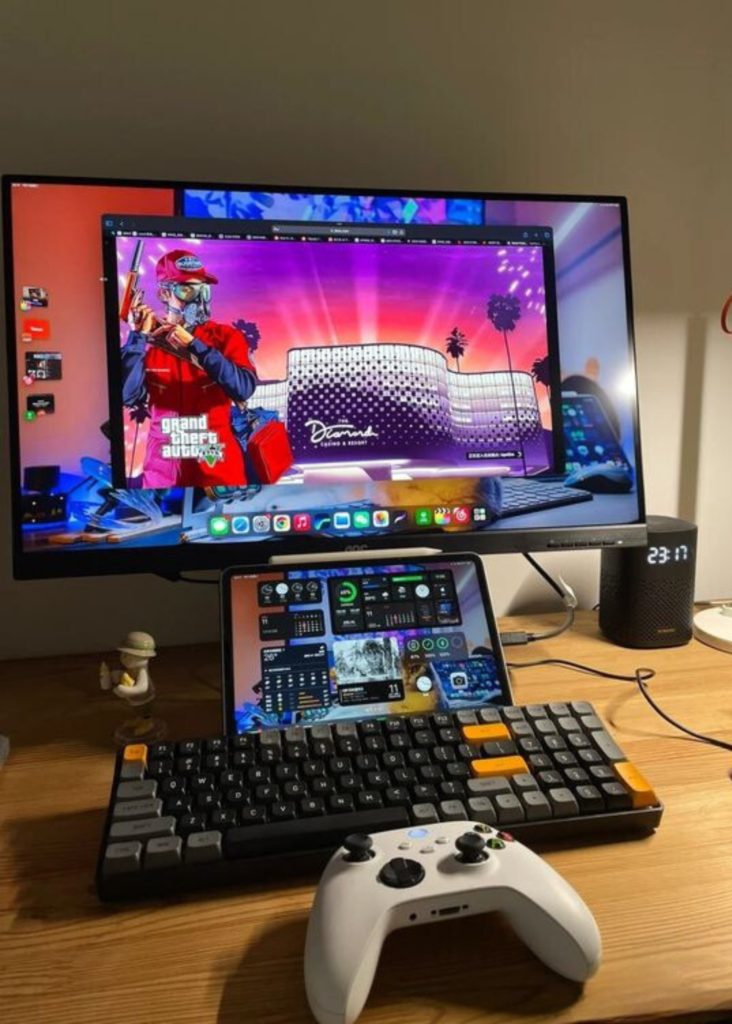 A gaming desk setup features a monitor displaying Grand Theft Auto V, a tablet with a dashboard, a mechanical keyboard, and a white game controller. A smart speaker showing the time, 23:17, and a small figurine are also present, with cables neatly arranged.